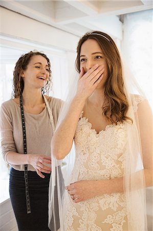 simsearch:6118-08971470,k - A young woman trying on a wedding dress, with lace overlay on the bodice and skirt, her hand covering her mouth, expressing surprise and delight. A dressmaker in the background. Photographie de stock - Premium Libres de Droits, Code: 6118-08971431