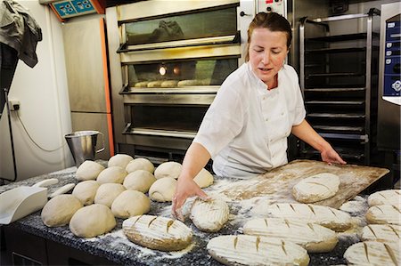 simsearch:6118-08971609,k - Woman standing at a table, placing freshly baked loaves of bread on a wooden board. Foto de stock - Royalty Free Premium, Número: 6118-08971571