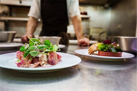 Close up of two plates of food in a kitchen, chef wearing apron standing in background. Stock Photo - Premium Royalty-Free, Code: 6118-08971593