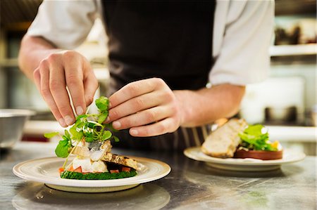 simsearch:6118-08971609,k - Close up of chef in kitchen adding salad garnish to a plate with grilled fish. Foto de stock - Royalty Free Premium, Número: 6118-08971599