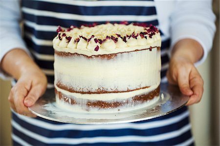 simsearch:6118-08971487,k - Close up of person wearing a blue and white stripy apron holding a cake decorated with cream and purple flower petals. Stockbilder - Premium RF Lizenzfrei, Bildnummer: 6118-08971512