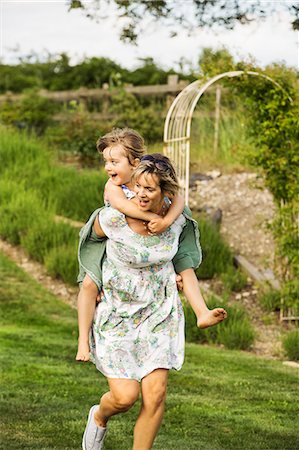 simsearch:6118-08971638,k - Woman in a sundress running across a lawn, giving a child a piggyback. Photographie de stock - Premium Libres de Droits, Code: 6118-08971660