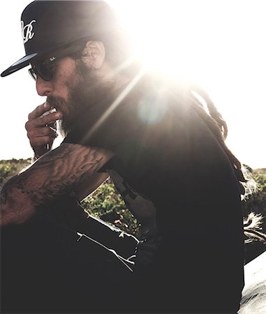 Side view of bearded man wearing baseball cap and sunglasses, smoking cigarette, tattooed arms, sunlight. Stock Photo - Premium Royalty-Free, Code: 6118-08991409