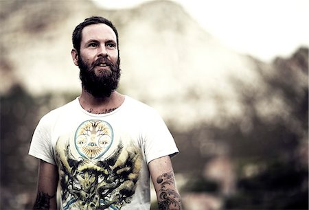 Portrait of bearded man wearing printed T-Shirt, tattoos on arms and chest, mountains in the background. Photographie de stock - Premium Libres de Droits, Code: 6118-08991412