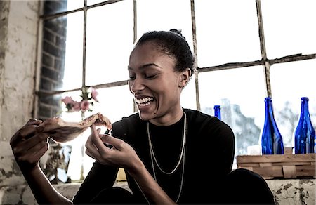 simsearch:6118-08991581,k - Smiling young woman sitting indoors by a window, eating slice of pizza. Stock Photo - Premium Royalty-Free, Code: 6118-08991578