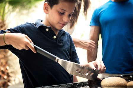 simsearch:6118-08991513,k - A boy holding tongs and turning food on a barbecue. Foto de stock - Royalty Free Premium, Número: 6118-08991508