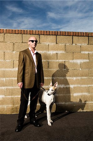 Man wearing a suit standing in front of a yellow brick wall, holding a white greyhound on a lead. Stock Photo - Premium Royalty-Free, Code: 6118-08928204