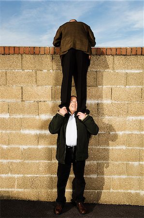 Rear view of man wearing a suit, standing on the shoulders of a man, climbing over yellow brick wall. Stock Photo - Premium Royalty-Free, Code: 6118-08928205