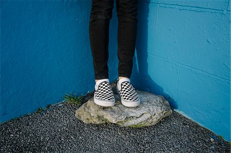 Teenage girl wearing black trousers and checkered canvas shoes, standing on rock against blue wall. Stock Photo - Premium Royalty-Free, Code: 6118-09059453