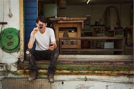 small business phone outside - A man in a teeshirt sitting on a step drinking from a cup, holding his smart phone. Stock Photo - Premium Royalty-Free, Code: 6118-09059756