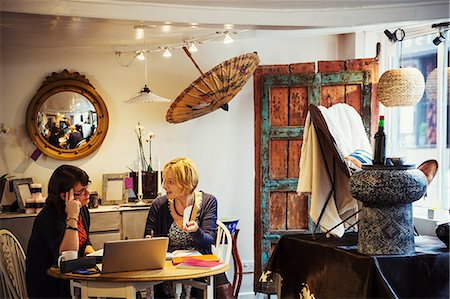 shopping laptop eyeglasses - Two women seated in a small retail outlet, a popup store, displays of retro and found objects, upcycled and renovated. Stock Photo - Premium Royalty-Free, Code: 6118-09059798