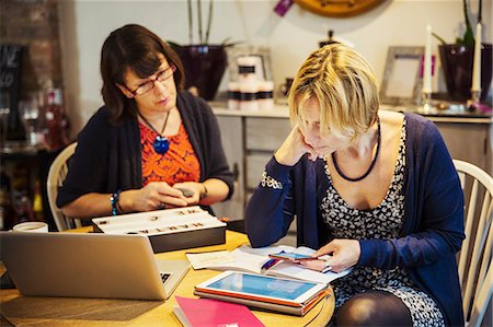 Two women seated at a table, one using a smart phone looking at a laptop screen. Stock Photo - Premium Royalty-Free, Code: 6118-09059799