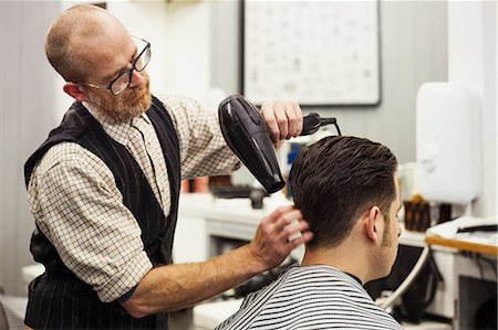 A customer sitting in the barber's chair, having his hair blow dried by a hairdresser and barber. Stock Photo - Premium Royalty-Free, Code: 6118-09059703