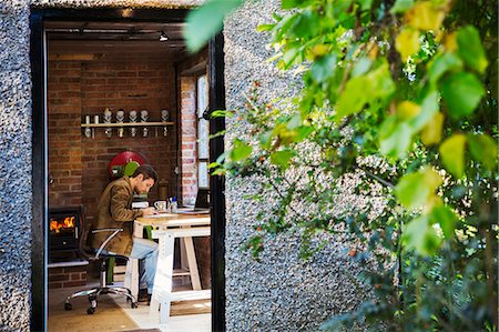 designer (high end design) - View through an open door, a designer seated in his leatherwork workshop at a desk, drawing. Woodburning stove with a glowing fire lit. Photographie de stock - Premium Libres de Droits, Code: 6118-09059817