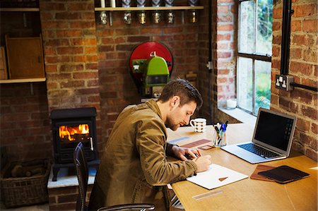 A designer seated in his leatherwork workshop, at a desk drawing on paper. A woodburning stove with a lit fire. Photographie de stock - Premium Libres de Droits, Code: 6118-09059816