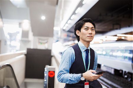 Businessman wearing blue shirt and vest standing on train station platform, holding mobile phone. Photographie de stock - Premium Libres de Droits, Code: 6118-09079184
