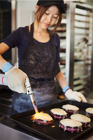 simsearch:649-03797461,k - Woman working in a bakery, wearing oven gloves, using blowtorch, melting cheese on sandwiches. Stockbilder - Premium RF Lizenzfrei, Bildnummer: 6118-09079248