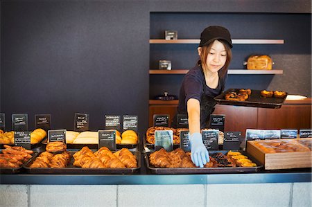 simsearch:6118-09079251,k - Woman working in a bakery, placing freshly baked croissants and cakes on large trays on a counter. Foto de stock - Sin royalties Premium, Código: 6118-09079252