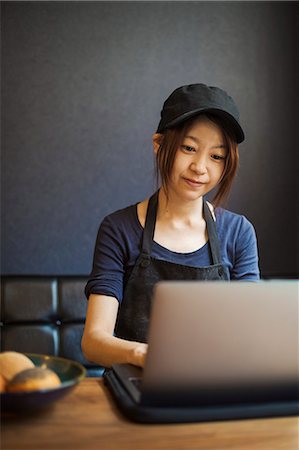 simsearch:6118-09079251,k - Woman working in a bakery, wearing baseball cap, sitting at table in front of laptop, typing. Foto de stock - Sin royalties Premium, Código: 6118-09079257