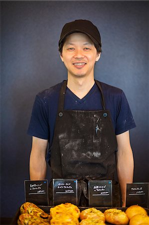 simsearch:6118-08660034,k - Smiling man wearing baseball cap and apron standing in a bakery, trays with freshly baked goods. Stock Photo - Premium Royalty-Free, Code: 6118-09079254