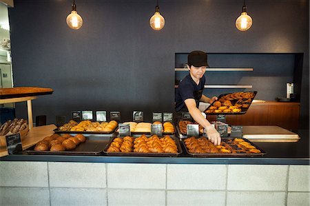 simsearch:6118-09079251,k - Man working in a bakery, placing freshly baked croissants and cakes on large trays on a counter. Foto de stock - Sin royalties Premium, Código: 6118-09079265