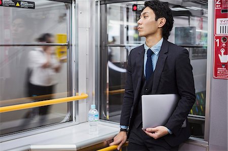 simsearch:640-01358423,k - Businessman wearing suit standing on a commuter train, holding laptop. Foto de stock - Royalty Free Premium, Número: 6118-09079213