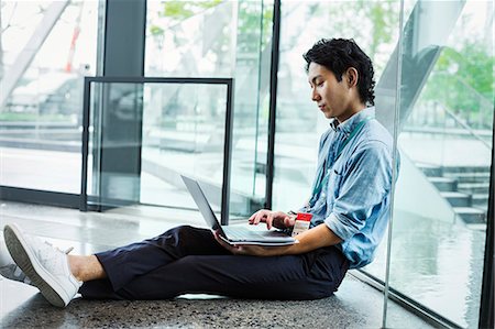 Businessman wearing blue shirt sitting on floor indoors, leaning against glass wall, working on laptop computer. Photographie de stock - Premium Libres de Droits, Code: 6118-09079222
