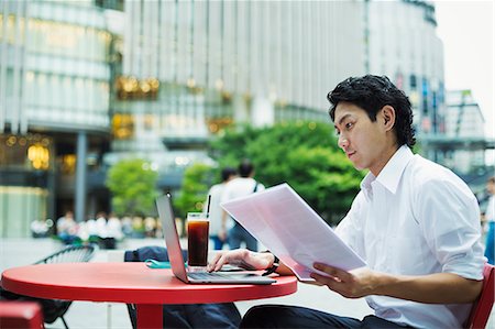 simsearch:6118-08351863,k - Businessman wearing white shirt sitting outdoors at red table, holding papers, working on laptop. Stock Photo - Premium Royalty-Free, Code: 6118-09079227