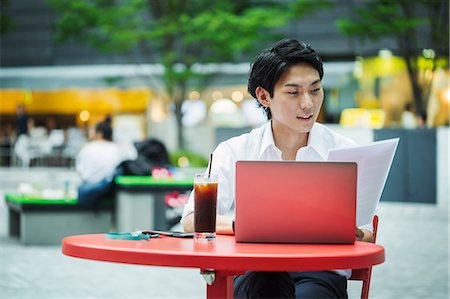 simsearch:6118-08351888,k - Businessman wearing white shirt sitting outdoors at red table, holding papers, working on laptop. Foto de stock - Royalty Free Premium, Número: 6118-09079228