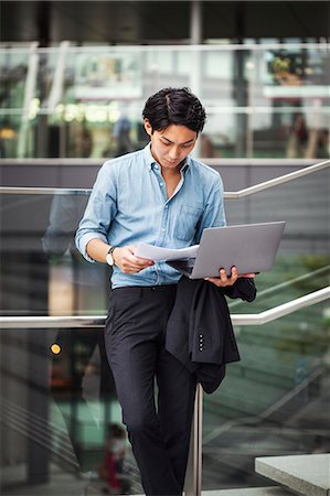 Businessman wearing blue shirt standing outdoors, holding laptop and papers, leaning against glass railing. Foto de stock - Sin royalties Premium, Código: 6118-09079238