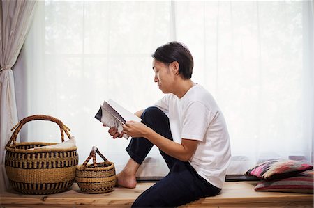 Man sitting indoors on a wooden bench with baskets, crossed legs, reading. Stock Photo - Premium Royalty-Free, Code: 6118-09079345