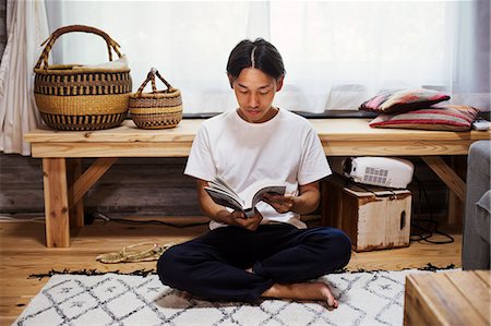 Man sitting indoors on a rug, leaning against wooden bench with baskets, crossed legs, reading. Stock Photo - Premium Royalty-Free, Code: 6118-09079346