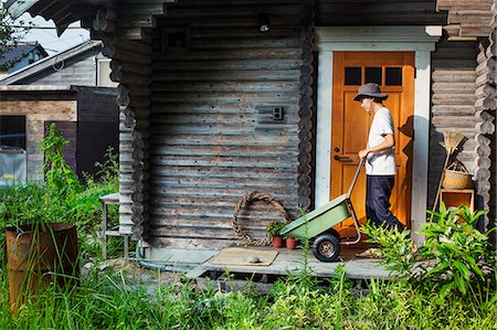 Man wearing hat standing outside wooden garden shed, pushing green wheelbarrow. Stock Photo - Premium Royalty-Free, Code: 6118-09079352