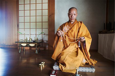 simsearch:6118-09079564,k - Buddhist monk with shaved head wearing golden robe kneeling indoors in a temple, holding mala. Fotografie stock - Premium Royalty-Free, Codice: 6118-09079367