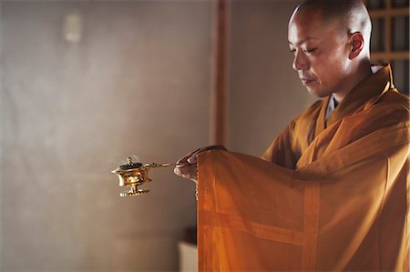 simsearch:6118-09079564,k - Side view of Buddhist monk with shaved head wearing golden robe kneeling indoors in a temple, holding incense burner. Fotografie stock - Premium Royalty-Free, Codice: 6118-09079365