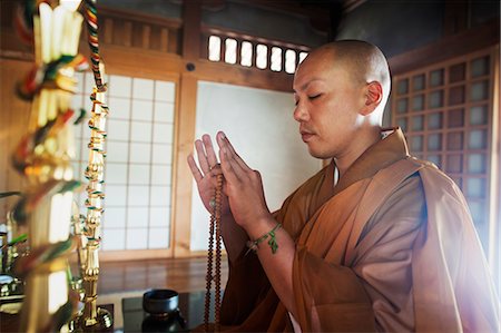 simsearch:6118-09079564,k - Side view of Buddhist monk with shaved head wearing golden robe kneeling indoors in a temple, holding mala, eyes closed. Fotografie stock - Premium Royalty-Free, Codice: 6118-09079366