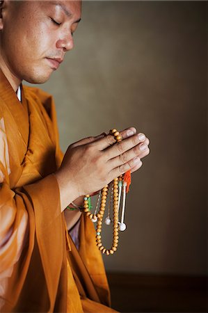 simsearch:6118-09200284,k - Side view of Buddhist monk with shaved head wearing golden robe kneeling indoors in a temple, holding mala, praying. Stock Photo - Premium Royalty-Free, Code: 6118-09079369
