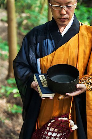 simsearch:6118-09079556,k - Buddhist monk with shaved head wearing black and yellow robe, standing outdoors, holding prayer book and singing bowl. Foto de stock - Sin royalties Premium, Código: 6118-09079383