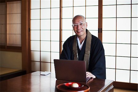 simsearch:6118-09079556,k - Buddhist monk with shaved head wearing black robe sitting indoors at a table, using laptop computer, smiling at camera. Foto de stock - Sin royalties Premium, Código: 6118-09079397