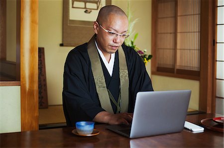 simsearch:6118-09079564,k - Buddhist monk with shaved head wearing black robe sitting indoors at a table, using laptop computer. Fotografie stock - Premium Royalty-Free, Codice: 6118-09079394