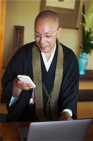 simsearch:6118-09079564,k - Buddhist monk with shaved head wearing black robe sitting indoors at a table in front of laptop computer, using mobile phone. Fotografie stock - Premium Royalty-Free, Codice: 6118-09079395
