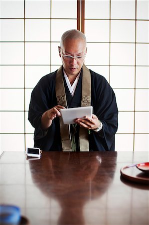simsearch:6118-09079556,k - Buddhist monk with shaved head wearing black robe sitting indoors at a table, using digital tablet. Foto de stock - Sin royalties Premium, Código: 6118-09079396