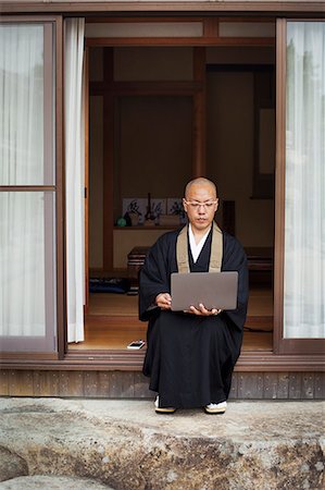 simsearch:6118-09079564,k - Buddhist monk with shaved head wearing black robe sitting outside temple, looking at laptop computer. Fotografie stock - Premium Royalty-Free, Codice: 6118-09079398