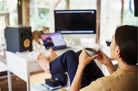 simsearch:6118-09079349,k - Man sitting indoors at a desk with computer, legs raised, barefoot, holding coffee mug. Stock Photo - Premium Royalty-Free, Code: 6118-09079333