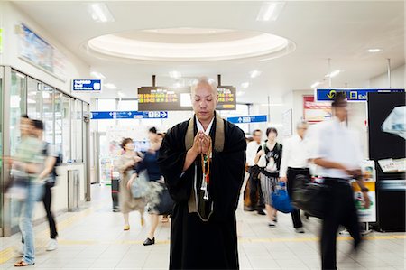 simsearch:6118-09200284,k - Buddhist monk with shaved head wearing black robe standing inside a train station, holding mala, people walking past. Stock Photo - Premium Royalty-Free, Code: 6118-09079403
