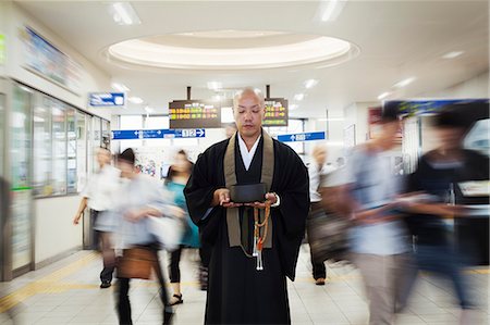 simsearch:6118-09200284,k - Buddhist monk with shaved head wearing black robe standing inside a train station, holding singing bowl and mala, people walking past. Stock Photo - Premium Royalty-Free, Code: 6118-09079402