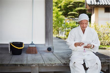 simsearch:6118-09079564,k - Buddhist monk wearing white robe and cap sitting on wooden floor outside a temple, using mobile phone. Fotografie stock - Premium Royalty-Free, Codice: 6118-09079407