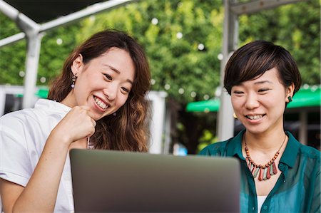 simsearch:6118-09079515,k - Two women with black hair wearing green and white shirt sitting in front of laptop at table in a street cafe, laughing. Stockbilder - Premium RF Lizenzfrei, Bildnummer: 6118-09079517