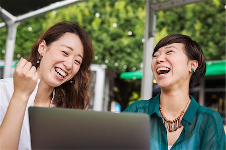 simsearch:6118-09079515,k - Two women with black hair wearing green and white shirt sitting in front of laptop at table in a street cafe, laughing. Stockbilder - Premium RF Lizenzfrei, Bildnummer: 6118-09079516