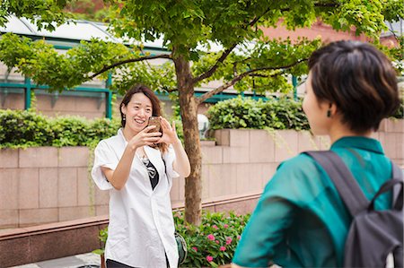 simsearch:6118-09079515,k - Two women with black hair wearing white and green shirt standing outdoors, taking picture with mobile phone, smiling. Stockbilder - Premium RF Lizenzfrei, Bildnummer: 6118-09079528